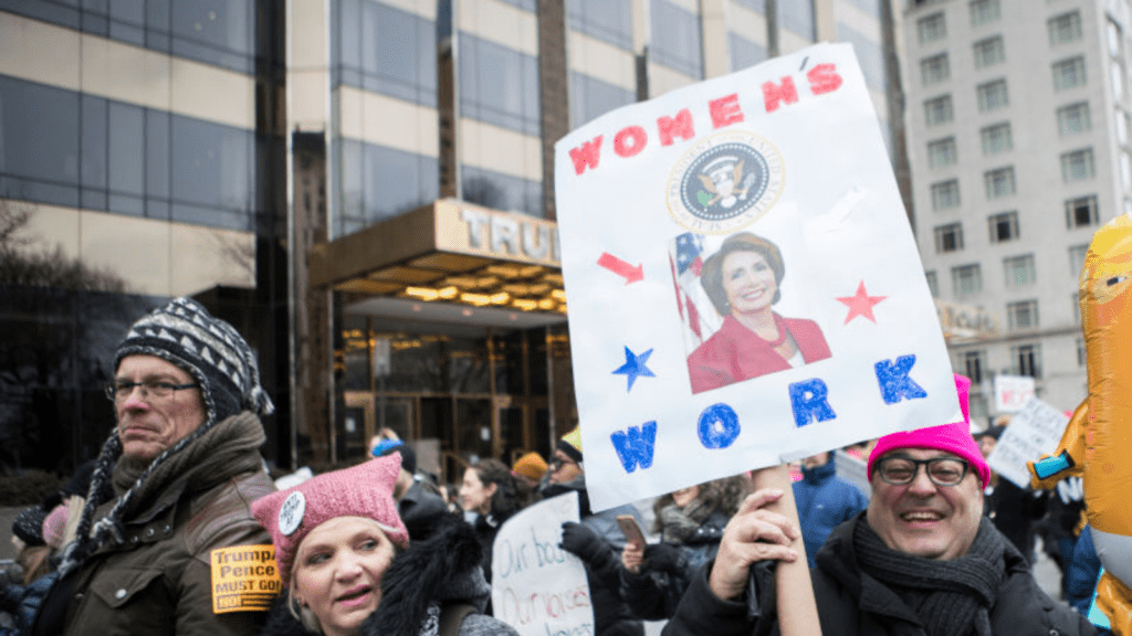 A marcher wearing a pink hat with a sign that says, "Women's Work" with a picture of Nancy Pelosi, Speaker of the United States House of Representatives and holding a balloon of Donald Trump in a diaper