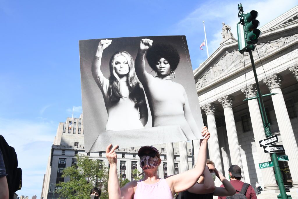 A protester holds a poster that shows an image of Gloria Steinem and Dorothy Pitman Hughes, each with a raised fist salute to demonstrate feminist solidarity.