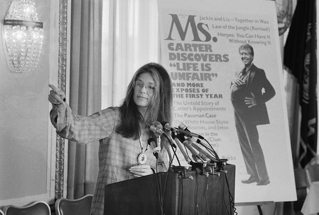 In a black and white photo, Gloria Steinem has long hair and glasses as she stands in front of a podium with multiple microphones. She is pointing outside the frame.