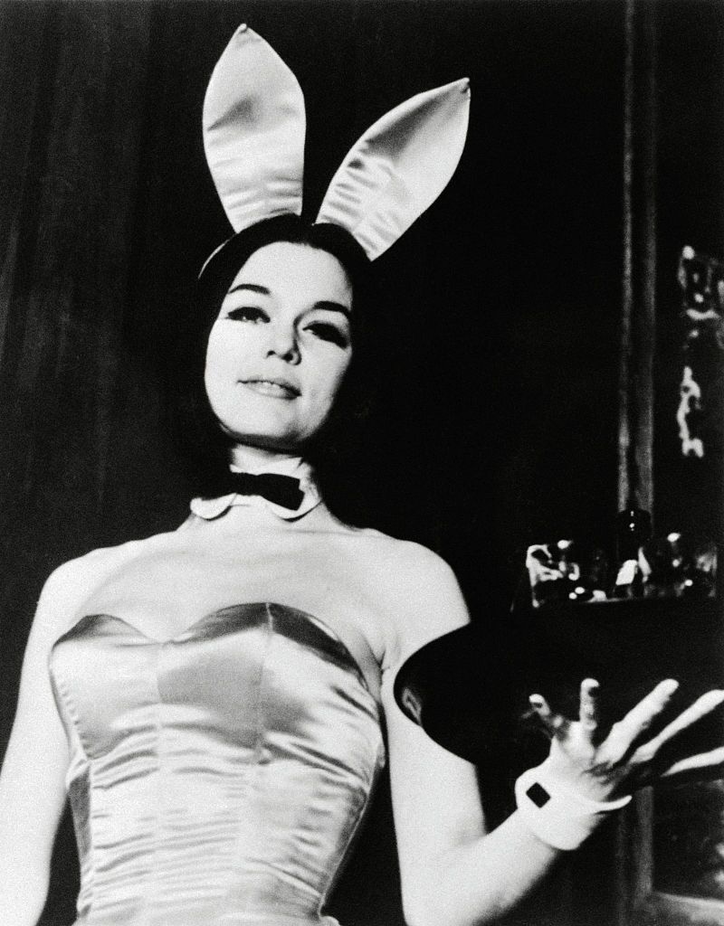 Young Gloria Steinem wears a Playboy bunny costume and holds a tray in this black and white photograph.