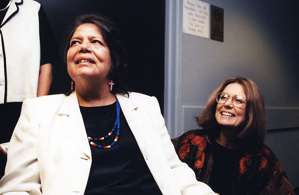 Steinem, wearing glasses and a red top, sits smiling behind Wilma Mankiller, a woman with black hair, beaded jewelry, and a white jacket.
