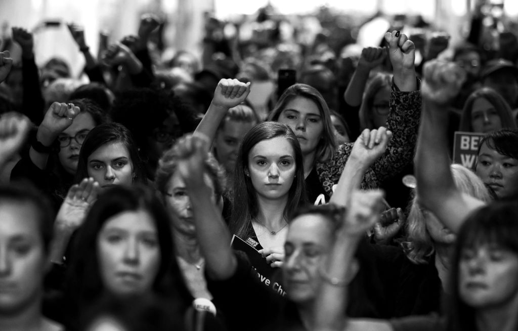 Čiernobiela fotografia ukazuje veľký dav demonštrantov, ktorí dvíhajú päste počas stretnutia #BelieveSurvivors proti nominácii Bretta Kavanaugha na najvyšší súd.