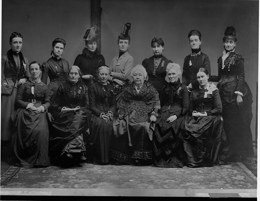 In a black-and-white image, a group of white women sit and stand for a photo. They wear traditional dresses from the 19th century. 