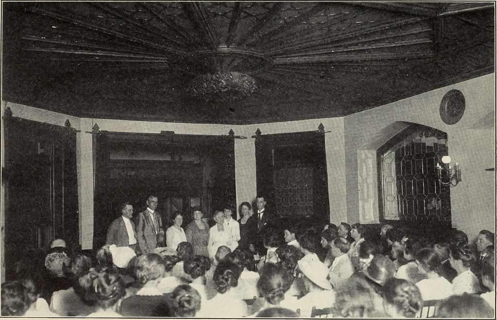 This black-and-white image displays a large, circular room with an intricate wood ceiling, where a group of men and women gathers for a meeting about women's suffrage. 