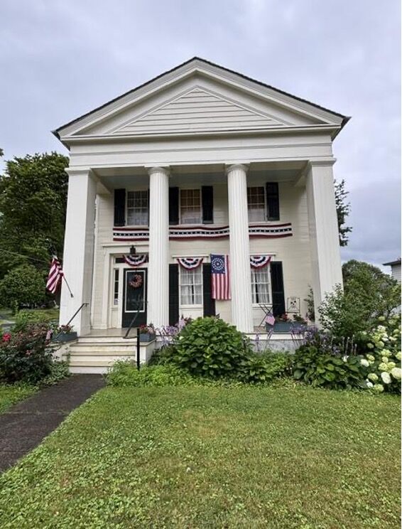 In the foreground of a cloudy sky stands a white two-story house with black shutters and cream pillars. American flags hang from the windows. The yard is green, and hydgrangeas sit out front. 